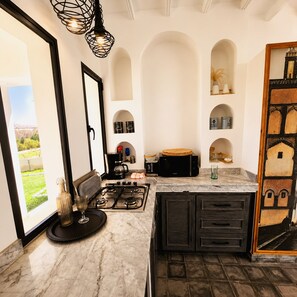 A modern kitchen with a gas stove, a coffee maker, and a view of a garden through a large window. - Palais Oriella (Essaouira)