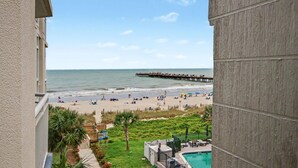 A beach view from a balcony with a pier extending into the ocean, a pool, and a grassy area with lounge chairs. - Oceanview 1BR Suite + Lazy River & Hot Tub (Myrtle Beach)