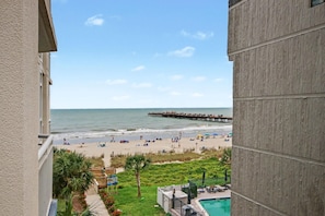 Blick auf einen Strand vom Balkon aus, mit einem Steg, der ins Meer führt, einem Pool und einer Rasenfläche mit Liegestühlen.