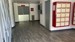 Un corridoio con una fila di cassette postali a destra, una porta con la scritta 'RDC' al centro e una parete grigia con una bocchetta di aerazione.