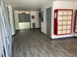 Ein Korridor mit einer Reihe von Briefkästen auf der rechten Seite, einer Tür mit der Aufschrift 'RDC' in der Mitte und einer grauen Wand mit einem Lüftungsgitter.