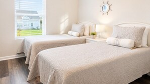 Um quarto com duas camas, uma mesa de cabeceira com luminária, um relógio na parede e uma janela com persianas.