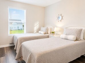 A bedroom with two beds, a nightstand with a lamp, a wall clock, and a window with blinds.