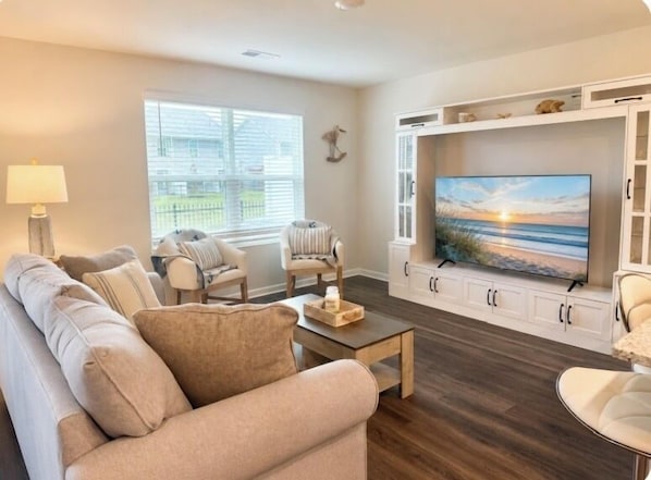 Un salon comprenant un canapé, des fauteuils, une table basse, un téléviseur posé sur un meuble blanc et une fenêtre avec des stores.