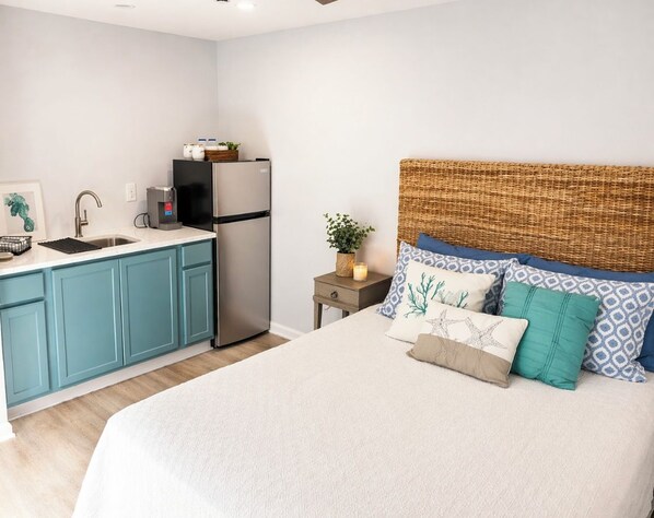 A bedroom with a bed, a nightstand, a small table, a sink, and a refrigerator.