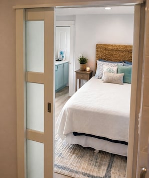 A bedroom with a bed, a bedside table, a nightstand, a lamp, and a door leading to a bathroom.