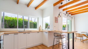 Una cocina moderna con armarios blancos, vigas de madera, ventanas grandes y una barra con taburetes.