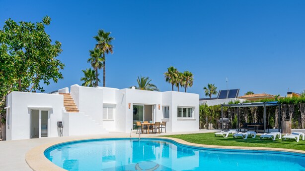 Un área moderna junto a la piscina con una piscina azul cristalina, paredes blancas y palmeras.