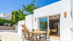 Un patio exterior moderno con una mesa y sillas de madera, un ventanal de vidrio que da a un espacio interior y una pared exterior blanca.