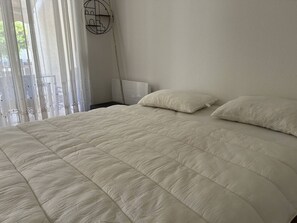 Ein ordentlich bezogenes Bett mit einer gesteppten Decke, ein Nachttisch mit Lampe und transparente Vorhänge, die natürliches Licht hereinlassen.