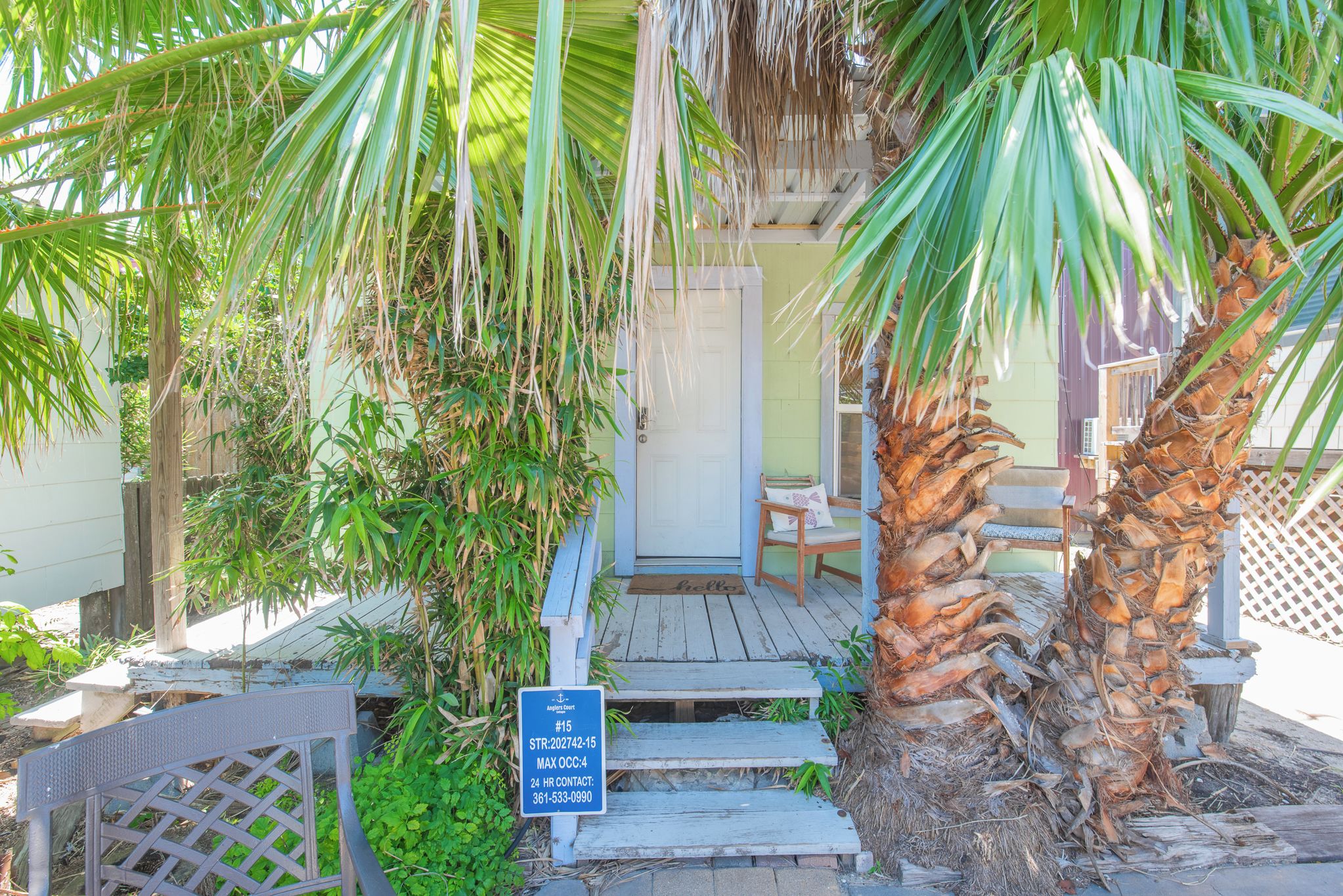 A wooden porch with steps leading to a door, surrounded by palm trees and a sign with contact information.