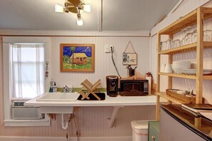 A kitchen with a white countertop, a microwave, a sink, and a window with curtains. - 1940s Old Town Beach Cabana Steps from Alister | Sleeps 2 (Port Aransas)