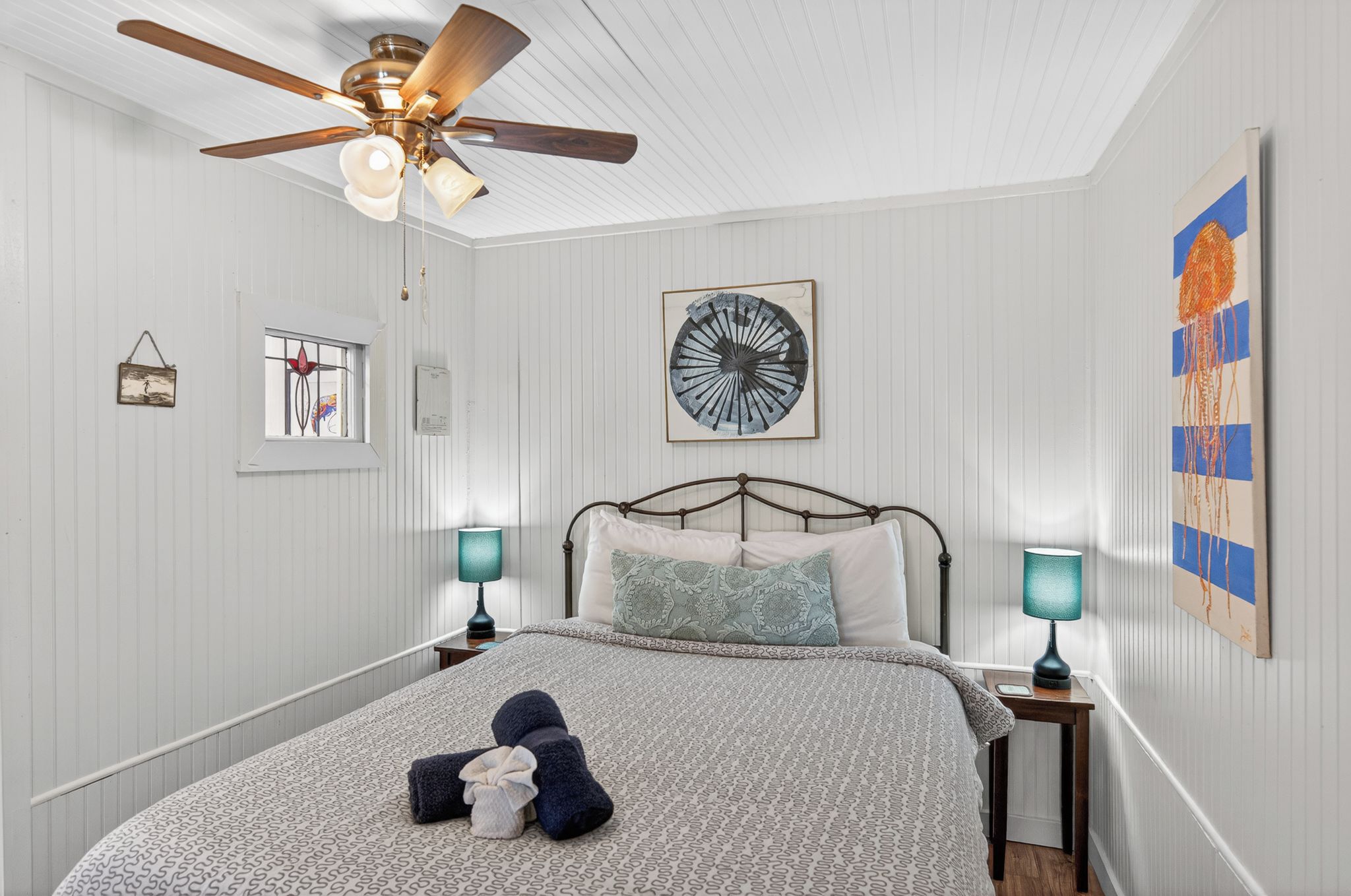 A bedroom with a bed, two bedside tables with lamps, a ceiling fan, a wall clock, and a large abstract painting.