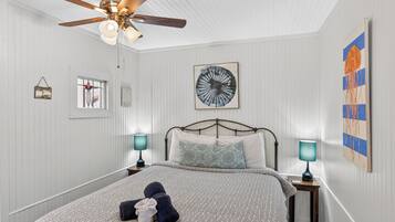 A bedroom with a bed, two bedside tables with lamps, a ceiling fan, a wall clock, and a large abstract painting.