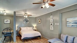 Un dormitorio con cama, ventilador de techo, escritorio, silla, sofá y cuadros en las paredes.
