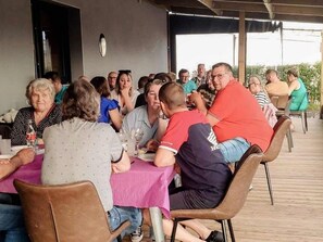 Un grupo de personas sentadas en mesas bajo un porche cubierto, evocando un ambiente de comedor comunitario.