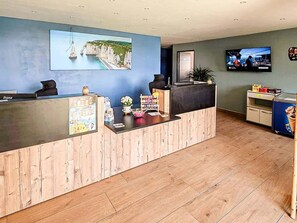 Un mostrador de recepción con detalles de madera, un cuadro enmarco de un paisaje costero, una pantalla de televisión y una máquina expendedora.