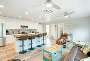 A modern kitchen with white cabinets, a central island with bar stools, a dining area with a wooden table and leather sofa, and a ceiling fan.