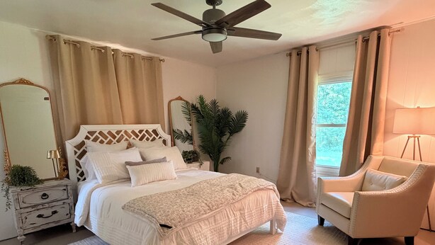 A bedroom with a bed, a chair, a ceiling fan, a window with curtains, a mirror, and a nightstand with a lamp. - Cozy Cottage on 9 tranquil acres in Wright City, Mo. Minutes to wedding venues! (Wright City)