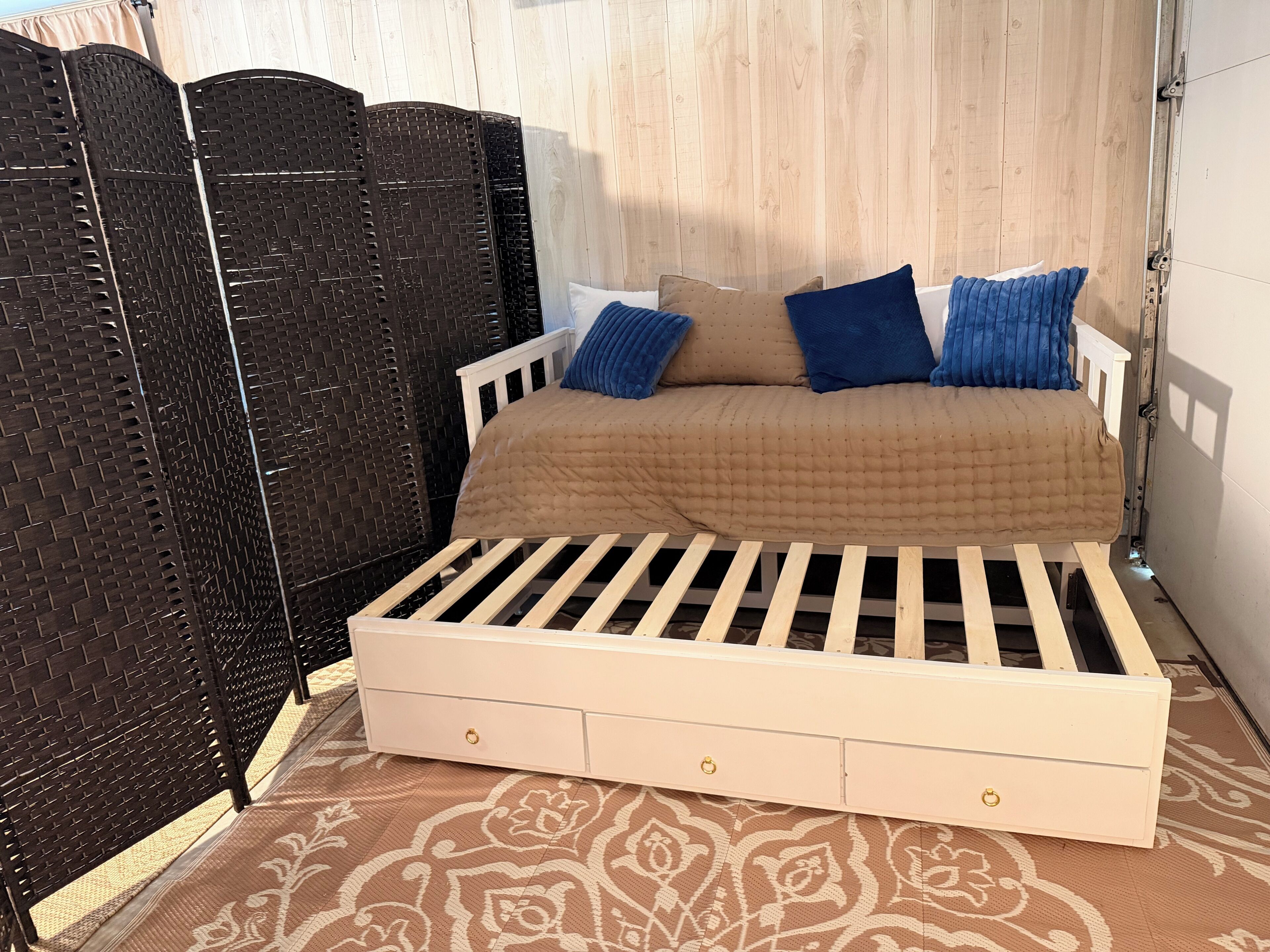 A single bed with a wooden frame and a quilted cover, accompanied by two pillows, in a room with a patterned floor and a wooden partition.