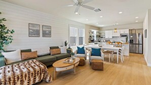 Amplia sala de estar con sofá, mesa de centro y reposapiés, un comedor con sillas, una cocina con electrodomésticos modernos y un ventilador de techo.
