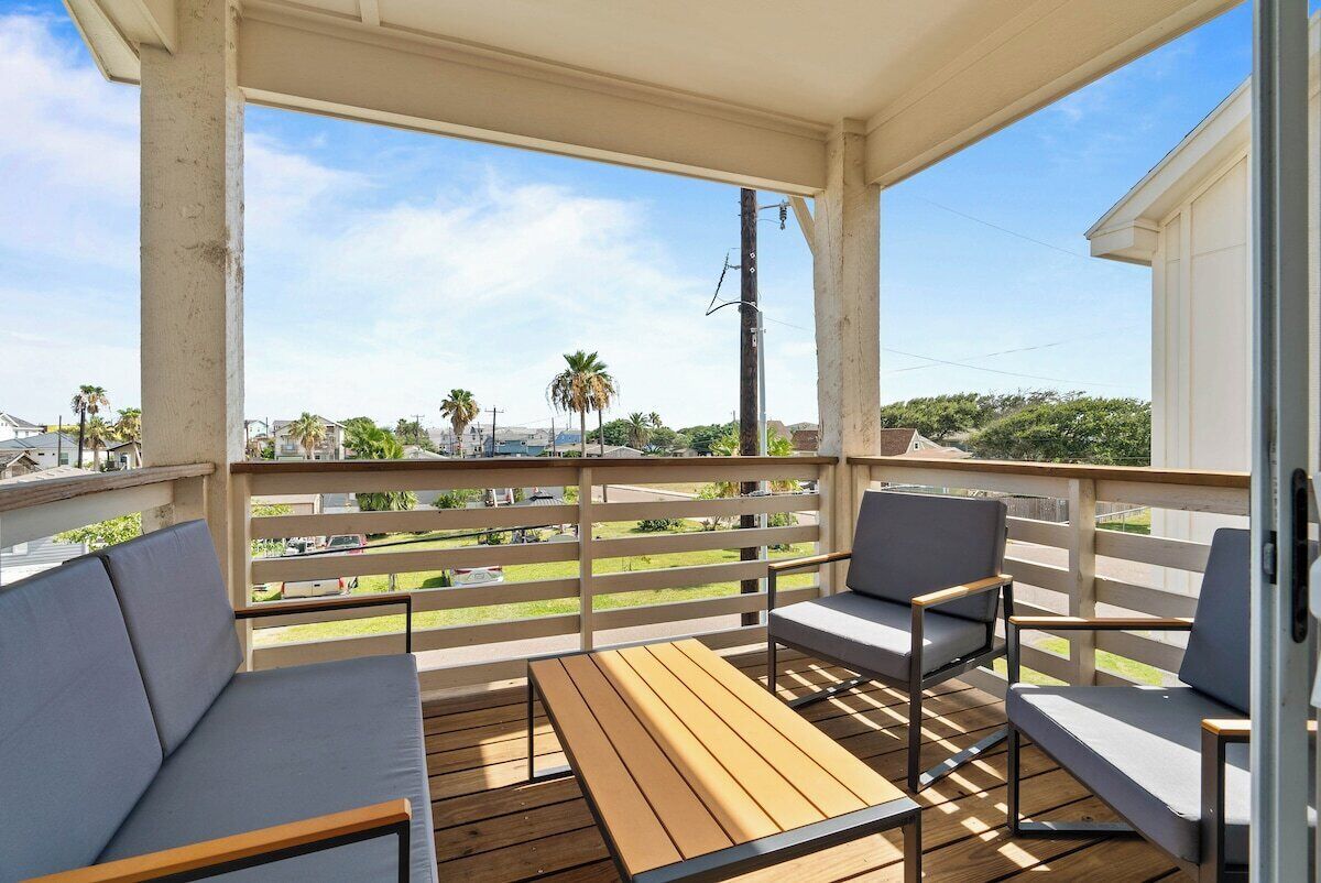 Een overdekt balkon met een houten tafel en twee stoelen, met uitzicht op een woongebied met palmbomen en huizen.