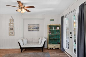 Ein Zimmer mit Deckenventilator, einer Couch, einem Bücherregal und einer Tür, die auf einen Balkon führt.