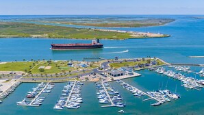 Vista aérea de uma marina com vários barcos atracados, um grande navio cargueiro ao fundo e uma pequena ilha com estruturas industriais.