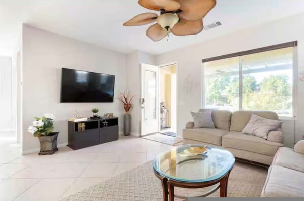 A living room with a ceiling fan, a flat-screen TV, a sofa, a glass coffee table, and a large window. - Country Club Home With Lakeside Views Golf (Indio)
