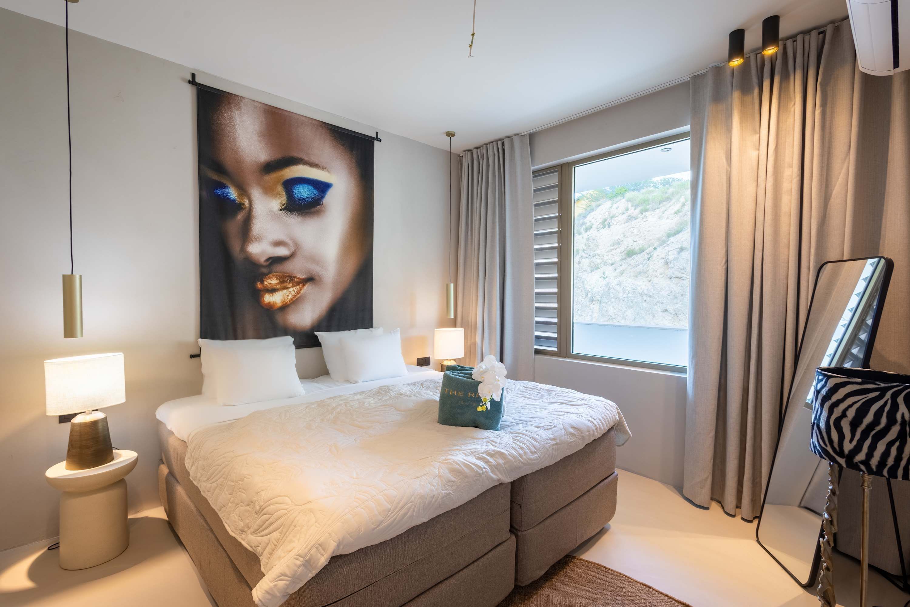 Quarto de hotel moderno com uma cama grande, duas luminárias de cabeceira, uma janela ampla com cortinas e uma obra de arte na parede.