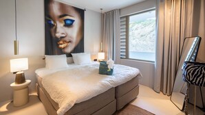 A modern hotel room with a large bed, two bedside lamps, a large window with curtains, and a wall-mounted artwork. - Aurel 13 Ocean View Penthouse at The Reef (Sint Michiel)