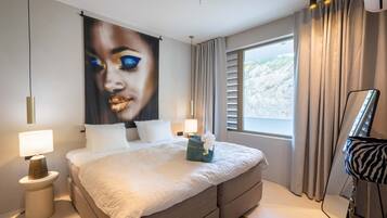 Quarto de hotel moderno com uma cama grande, duas luminárias de cabeceira, uma janela ampla com cortinas e uma obra de arte na parede.