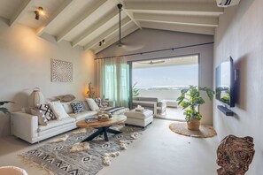 Ein modernes Wohnzimmer mit einem weißen Sofa, einem Couchtisch, einem an der Wand montierten Fernseher und einem großen Fenster mit Blick.