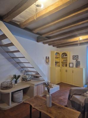 Un intérieur rustique avec des poutres apparentes, un escalier, un buffet jaune aux portes vitrées et une table en bois sur laquelle est posé un vase.