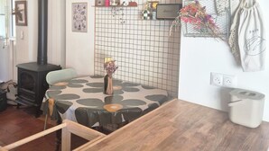 Une cuisine rustique avec un poêle à bois, un dosseret carrelé, une étagère suspendue ornée d’objets décoratifs et une table recouverte d’une nappe à motifs.