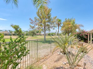 A fenced outdoor area with a variety of trees and shrubs, a clear sky, and a distant view of houses. - Pools Golf and Tennis Home (Indio)