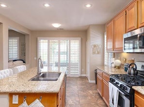 A kitchen with a granite countertop, stainless steel appliances, wooden cabinets, and a large window with blinds. - Pools Golf and Tennis Home (Indio)