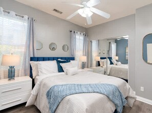 A bedroom with a bed, two bedside tables, a ceiling fan, and a large mirror. - Pools Golf and Tennis Home (Indio)