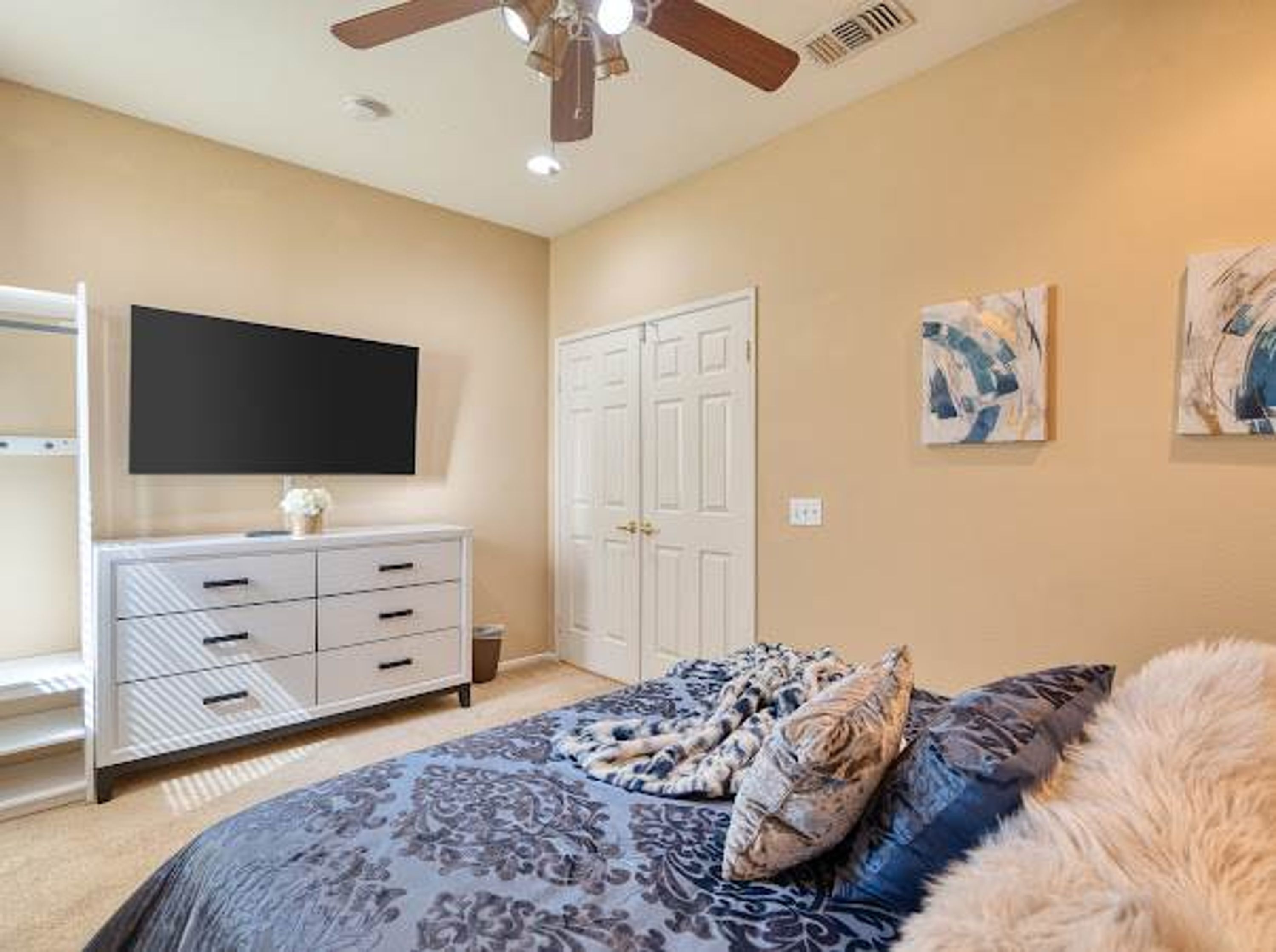 A bedroom with a bed, a ceiling fan, a TV, a wardrobe, and artwork on the wall.