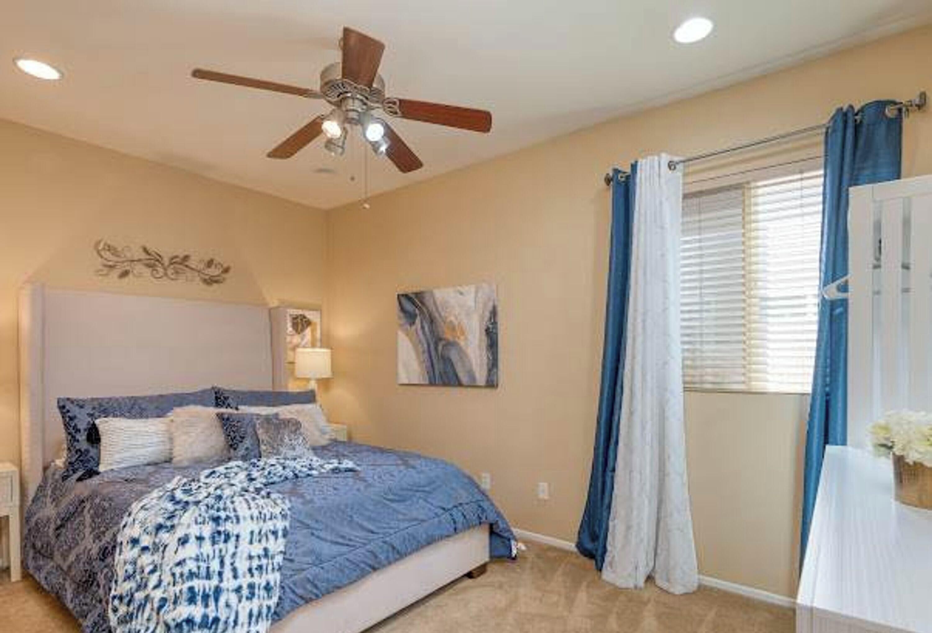 Une chambre à coucher comprenant un lit, un ventilateur de plafond, une fenêtre avec des rideaux et une œuvre d’art accrochée au mur.