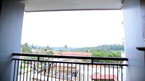 A balcony with a metal railing, a view of a street with buildings, and a distant landscape with trees and hills. - Aldur Residency (Chikkamagaluru)