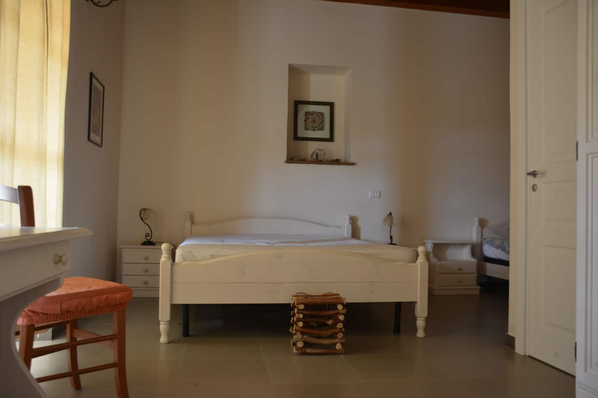 A bedroom with a bed, a chair, a bedside table, a nightstand, a wall-mounted shelf, and a door.