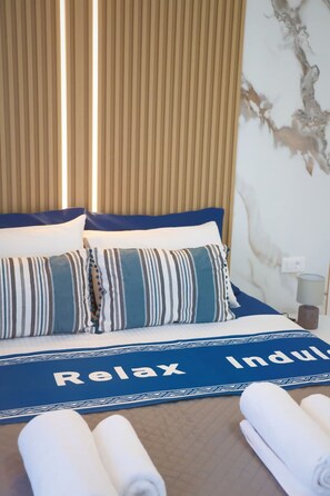 Ein Hotelzimmer mit einem Bett, Kissen, einer blauen Bettdecke mit dem Aufdruck 'Relax Indulge' und einer Wand mit Marmor-Optik sowie einem Fenster mit Jalousien.