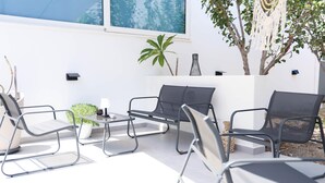 Un área de estar al aire libre con sillas de metal y una mesa, rodeada de plantas en macetas y un árbol.