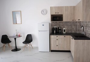 Eine kompakte Küche mit Kühlschrank, Mikrowelle, Kaffeemaschine und Spüle. Ein kleiner Esstisch mit zwei Stühlen und einer Uhr an der Wand.