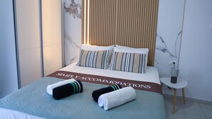 A neatly made bed with striped pillows, a 'SIMPLE ACCOMMODATIONS' sign, and two towels on the bed. - Apartment Simply 205 - Ammoudara Sandy Retreat (Malevizi)