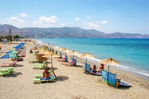 Ein Strand mit Reihen bunter Sonnenschirme und Liegestühle, ruhiges Meer und Berge im Hintergrund.