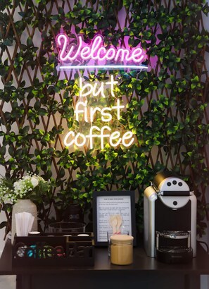 Eine Neonreklame mit der Aufschrift 'Welcome but first coffee' vor einem Grünpflanzenhintergrund.