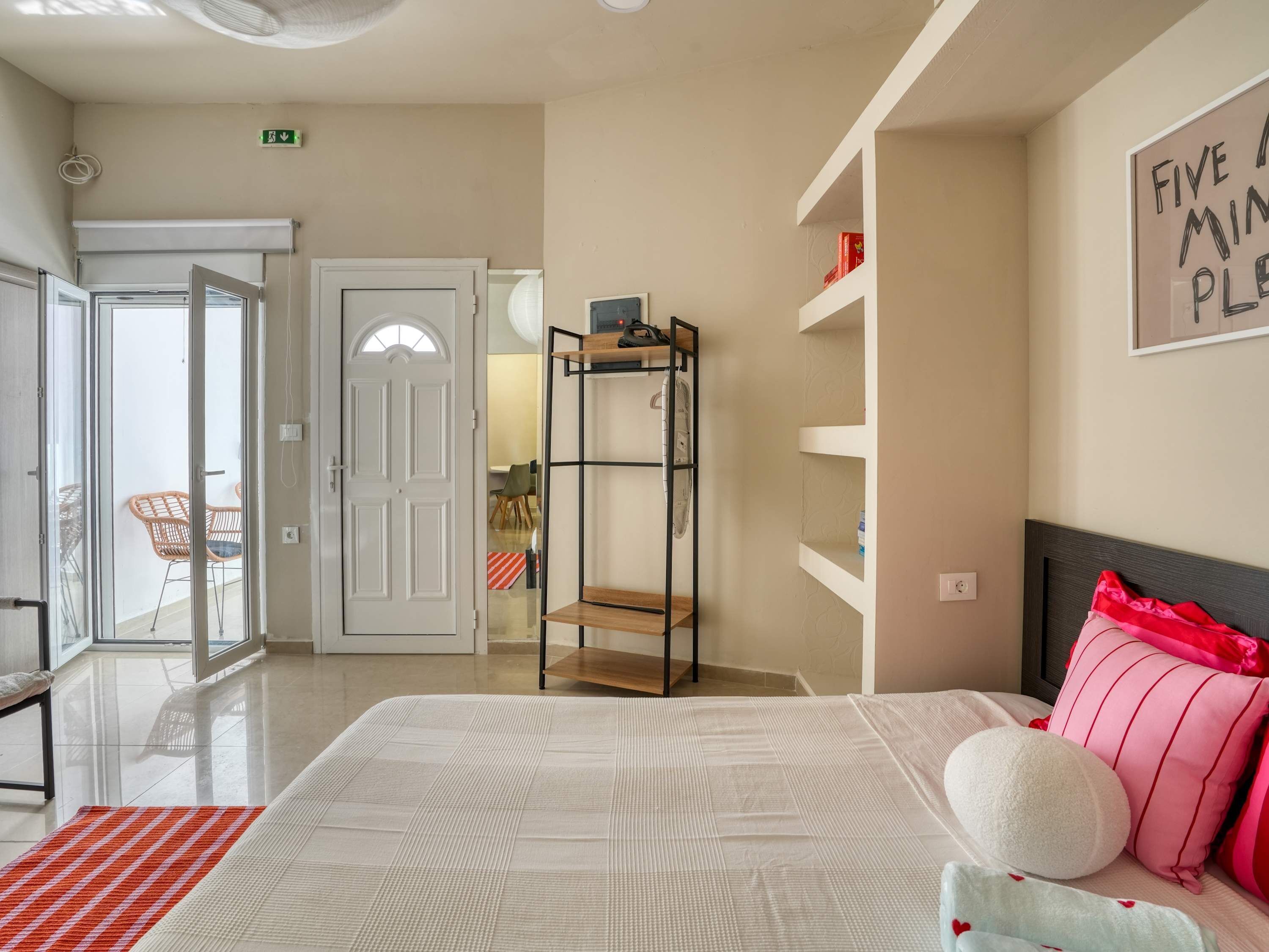Quarto com cama, mesa de cabeceira, prateleira na parede e porta para uma varanda.