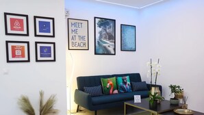 A modern living room with a dark blue sofa, framed pictures on the wall, a lamp, and a coffee table with a plant. - Apartment Simply 101 - Ammoudara Sandy Retreat (Malevizi)